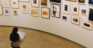 "A focused individual is seen examining a list of sold masterpieces on a gallery wall, beneath the prominent inscription 'Art Sells Best.' The list includes titles and prices, indicating the successful sales of various artworks, and the person's intrigued expression suggests they are analyzing the trends and popularity of different art pieces in the market."